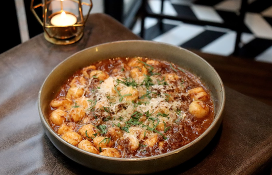 A plate of parmesan and parsley covered gnocchi with a beef cheek ragu on a leather covered table with a tea light candle in the background.