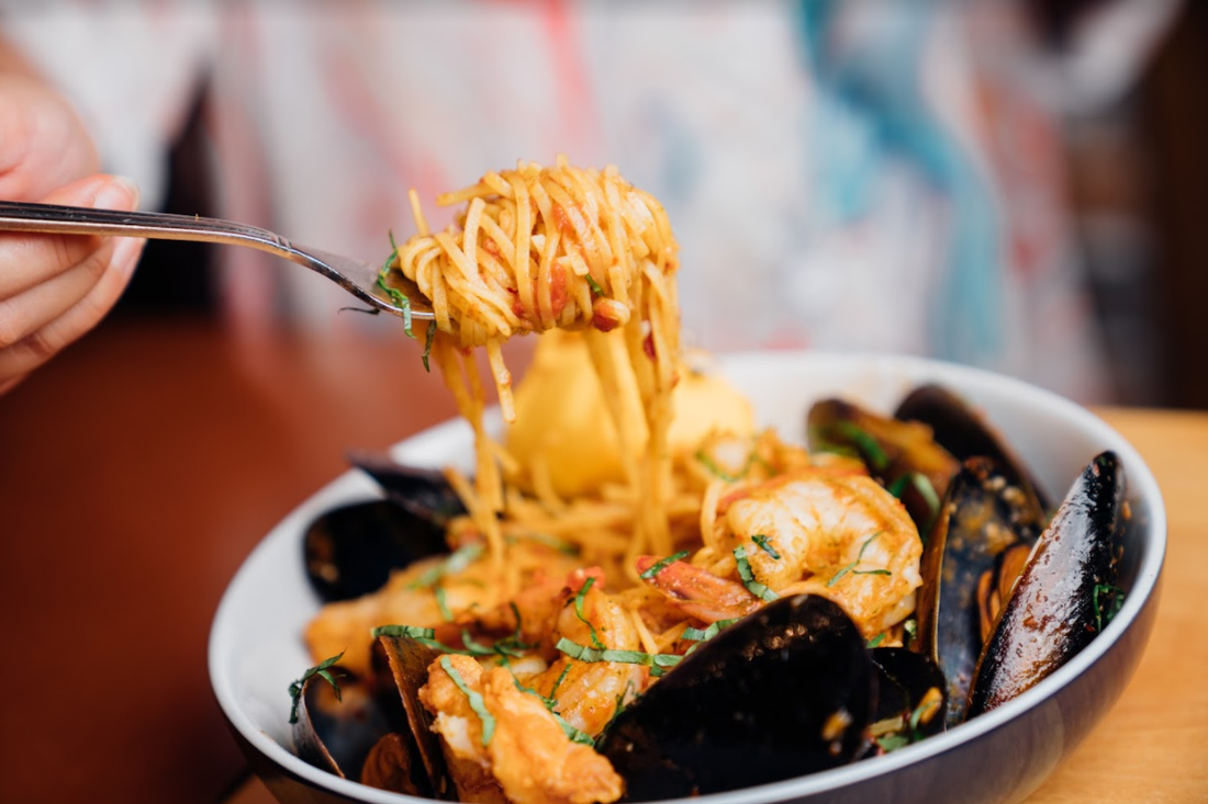 A forkful of pasta being held above a bowl of seafood pasta with mussels and prawns in a red sauce.
