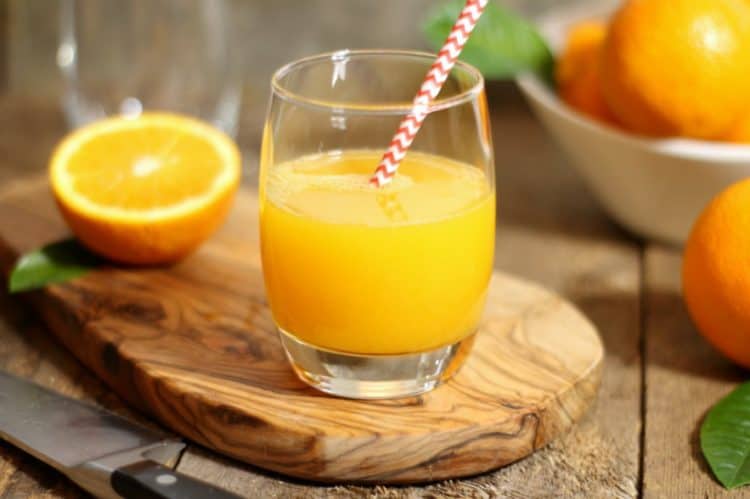 A glass of freshly squeezed orange juice with a paper straw, half an orange all on a wooden board.